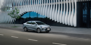 Chevrolet Onix Plus na cor Prata Shark em avenida com fachada arquitetônica de linhas onduladas ao fundo.