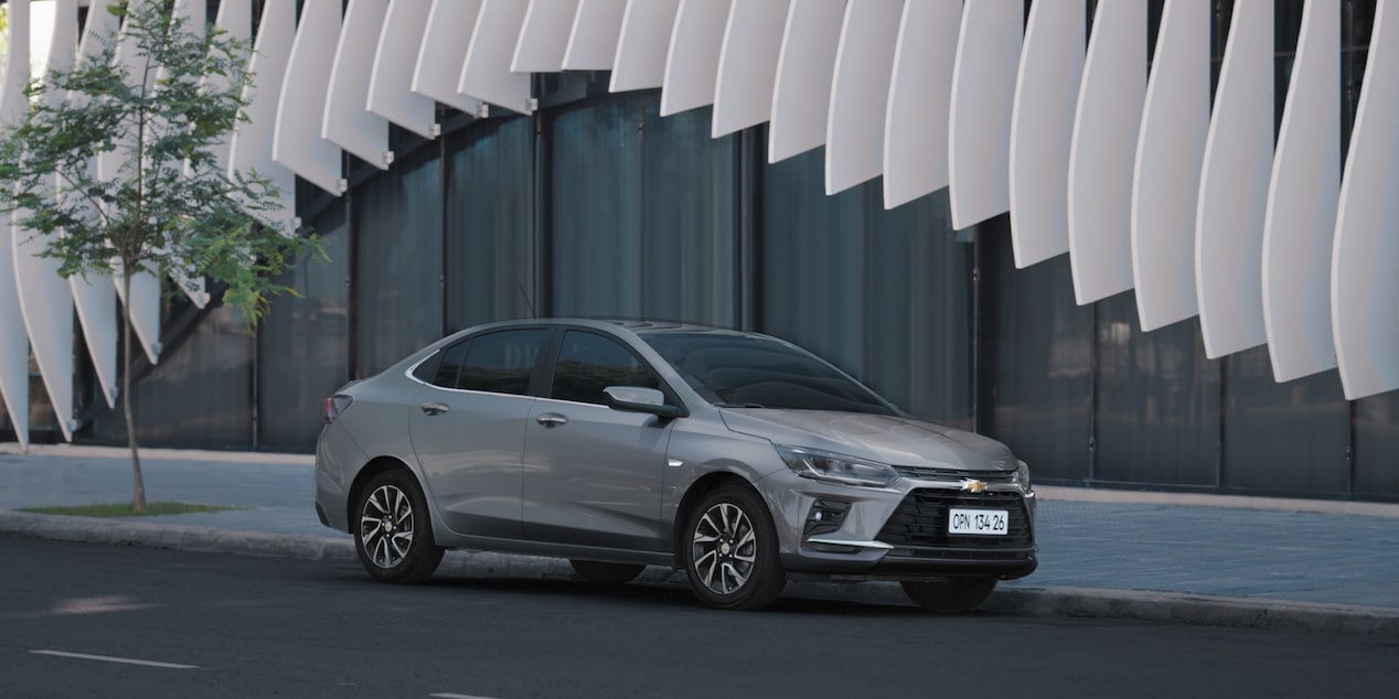 Chevrolet Onix Plus na cor Prata Shark estacionada com fachada arquitetônica de linhas onduladas ao fundo.