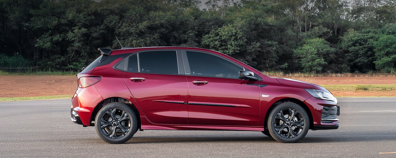 Chevrolet Onix HB na versão RS em fotografia ambientada. O veículo é visto lateralmente e ilustra a página de acessórios originais Chevrolet dedicada a ele próprio. No quadrante superior esquerdo, o texto: Onix - Aparência.