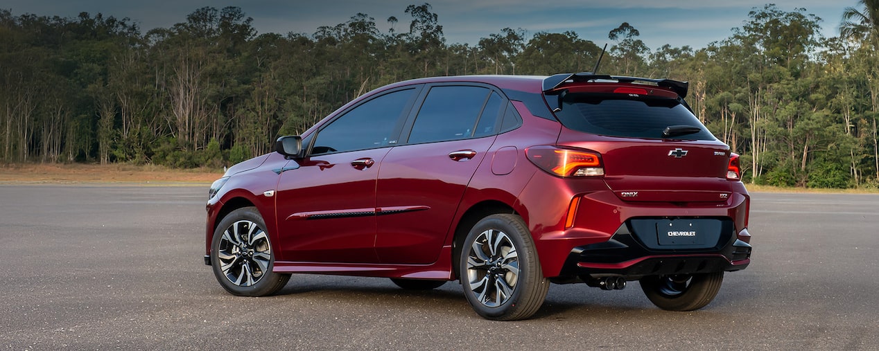 Masthead versão desktop da página de Pacotes, dentro de acessórios originais Chevrolet, para o Onix, que aparece em foto ambientada, estacionado diante de uma grande parede de árvores verdinhas. 