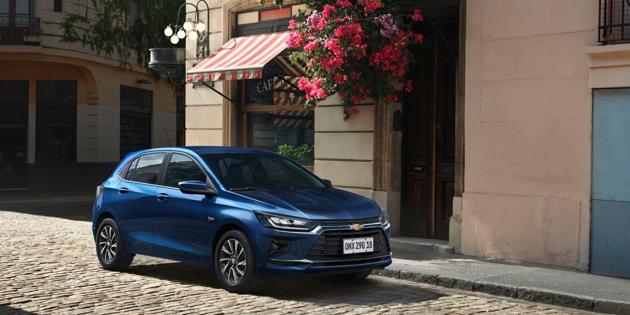 Chevrolet Onix hatch azul estacionado em rua de paralelepípedo, carro compacto moderno com design elegante, faróis em LED e cenário urbano charmoso.