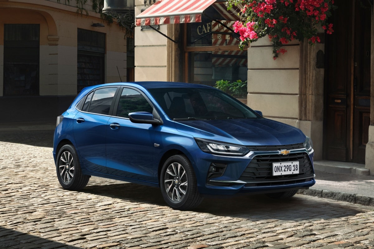 Chevrolet Onix hatch azul estacionado em rua de paralelepípedo, carro compacto moderno com design elegante, faróis em LED e cenário urbano charmoso.