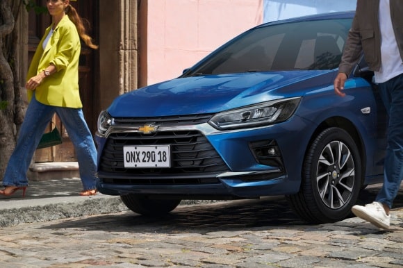 Chevrolet Onix hatch azul em rua de paralelepípedo, destaque para frente do carro com faróis LED e design moderno em cenário urbano.