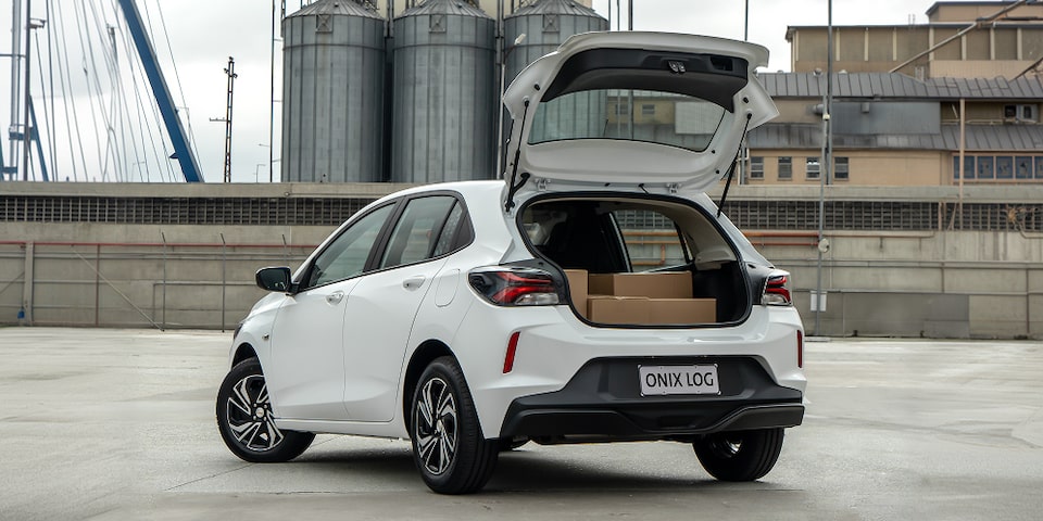 Chevrolet Onix Log branco com porta-malas aberto, exibindo capacidade de carga com caixas, em ambiente industrial urbano.