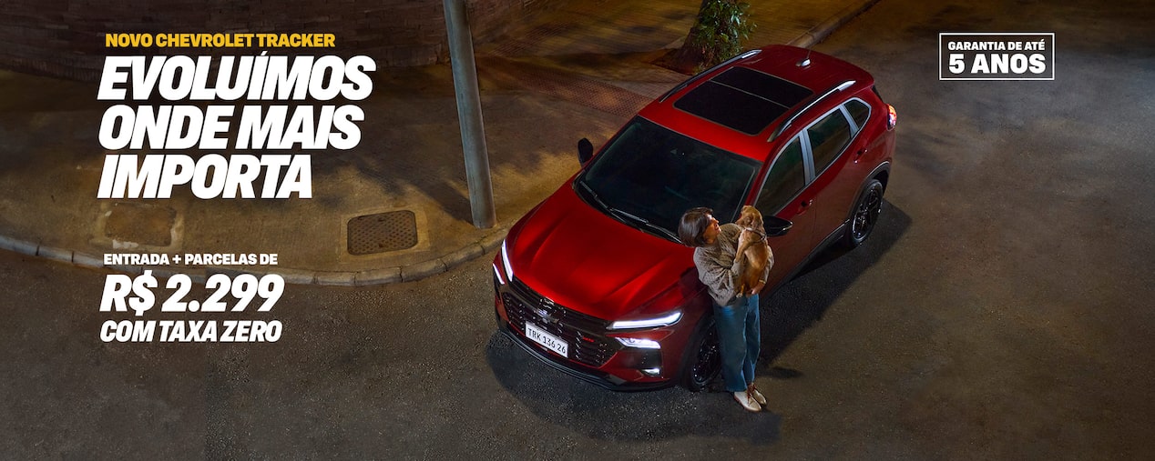 Novo Chevrolet Tracker, cor vermelha, visto de cima em uma rua à noite. Uma mulher segura um cão pequeno ao lado do carro. Texto promocional: Evoluímos onde mais importa. Taxa zero e parcelas de apenas R$ 2.299 com 5 anos de garantia. Selo Garantia de até 5 anos no canto superior direito.