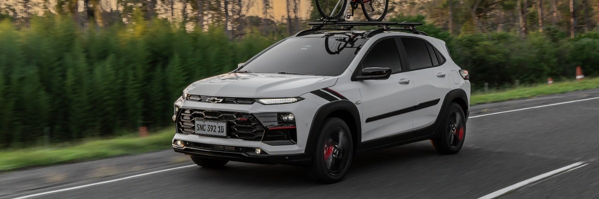 Chevrolet Sonic em movimento em uma estrada. Ao fundo vemos alguns arbustos e árvores. Em cima do veículo há uma bicicleta fixada no rack de teto.