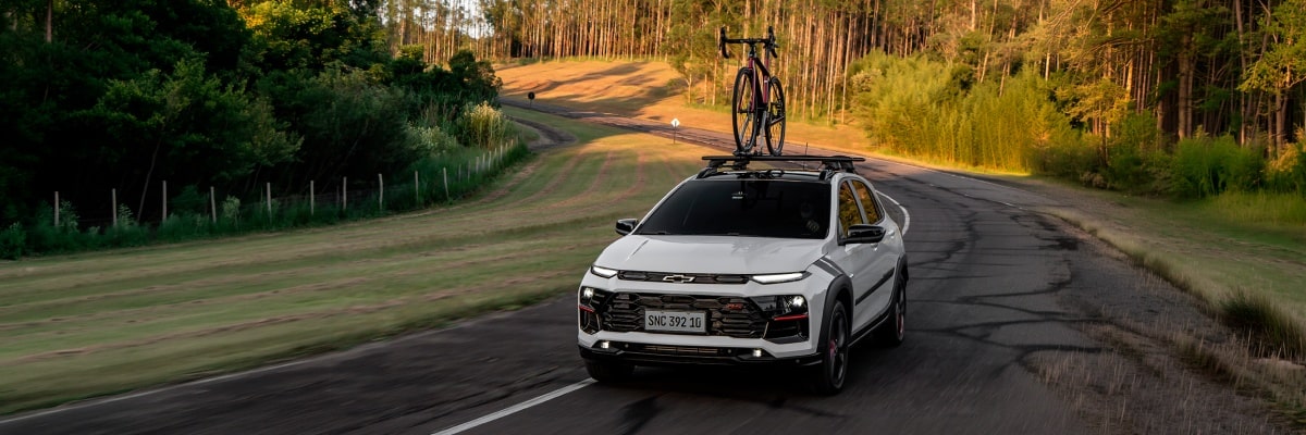 Chevrolet Sonic em movimento em uma estrada. Ao fundo vemos alguns arbustos e árvores. Em cima do veículo há uma bicicleta fixada no rack de teto.
