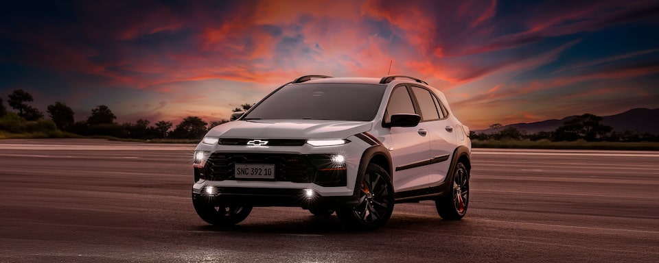 Chevrolet Sonic estacionado em uma área externa. O céu está com uma coloração avermelhada de fim de tarde e o veículo está com os faróis ligados. Ao fundo, algumas árvores.