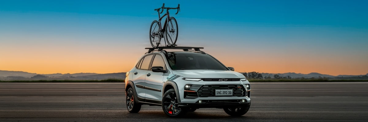 Chevrolet Sonic estacionado em uma área externa. O céu está com uma coloração levemente avermelhada de fim de tarde e o veículo está com os faróis ligados. Há uma bicicleta fixada no rack de teto do veículo.