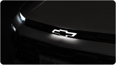 Parte frontal do Chevrolet Sonic com destaque para o logo da Chevrolet e lanternas acesas. Todo o ambiente ao redor está escuro.