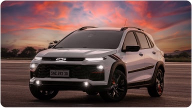 Chevrolet Sonic estacionado em uma área externa. O céu está com uma coloração avermelhada de fim de tarde e o veículo está com os faróis ligados. Ao fundo, algumas árvores.