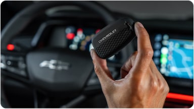 Temos no primeiro plano uma mão segurando a chave de presença preta do Chevrolet Sonic. Em segundo plano, temos o volante e o monitor do veículo.