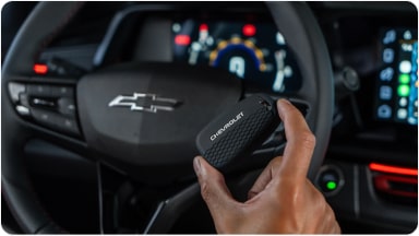 Temos no primeiro plano uma mão segurando a chave de presença preta do Chevrolet Sonic. Em segundo plano, temos o volante e o monitor do veículo.