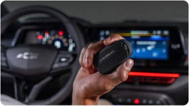 Temos no primeiro plano uma mão segurando a chave de presença preta do Chevrolet Sonic. Em segundo plano, temos o volante e o monitor do veículo.