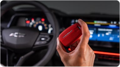 Temos no primeiro plano uma mão segurando a chave de presença vermelha do Chevrolet Sonic. Em segundo plano, temos o volante e o monitor do veículo.