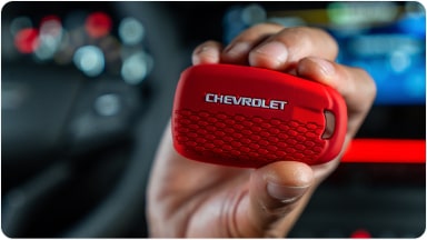 Temos no primeiro plano uma mão segurando a chave de presença vermelha do Chevrolet Sonic. Em segundo plano, temos o volante e o monitor do veículo.