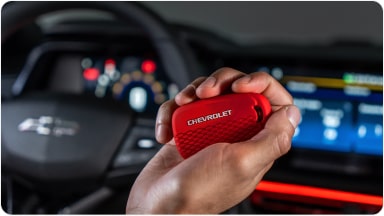 Temos no primeiro plano uma mão segurando a chave de presença vermelha do Chevrolet Sonic. Em segundo plano, temos o volante e o monitor do veículo.