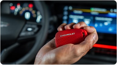 Temos no primeiro plano uma mão segurando a chave de presença vermelha do Chevrolet Sonic. Em segundo plano, temos o volante e o monitor do veículo.