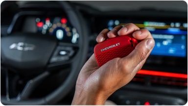 Temos no primeiro plano uma mão segurando a chave de presença vermelha do Chevrolet Sonic. Em segundo plano, temos o volante e o monitor do veículo.
