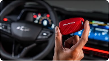 Temos no primeiro plano uma mão segurando a chave de presença vermelha do Chevrolet Sonic. Em segundo plano, temos o volante e o monitor do veículo.