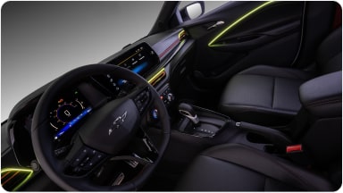 Visão interna do veículo. Na imagem, temos todo o painel, volante e parte dos bancos do motorista e passageiro. Alguns detalhes em led amarelo na porta e do lado do volante estão acesos dentro do veículo.