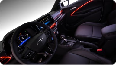 Visão interna do veículo. Na imagem, temos todo o painel, volante e parte dos bancos do motorista e passageiro. Alguns detalhes em led vermelho na porta e do lado do volante estão acesos dentro do veículo.
