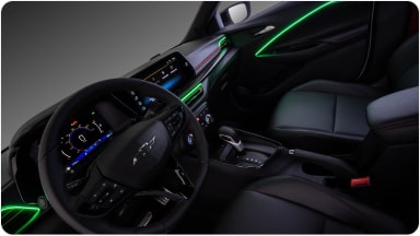 Visão interna do veículo. Na imagem, temos todo o painel, volante e parte dos bancos do motorista e passageiro. Alguns detalhes em led verde na porta e do lado do volante estão acesos dentro do veículo.
