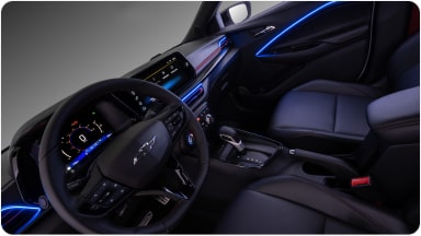 Visão interna do veículo. Na imagem, temos todo o painel, volante e parte dos bancos do motorista e passageiro. Alguns detalhes em led azul na porta e do lado do volante estão acesos dentro do veículo.