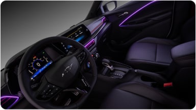 Visão interna do veículo. Na imagem, temos todo o painel, volante e parte dos bancos do motorista e passageiro. Alguns detalhes em led roxo na porta e do lado do volante estão acesos dentro do veículo.