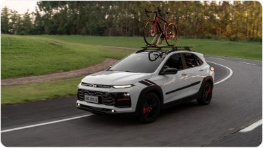Chevrolet Sonic em movimento em uma estrada. Ao fundo vemos alguns arbustos e árvores. Em cima do veículo há uma bicicleta fixada no rack de teto.