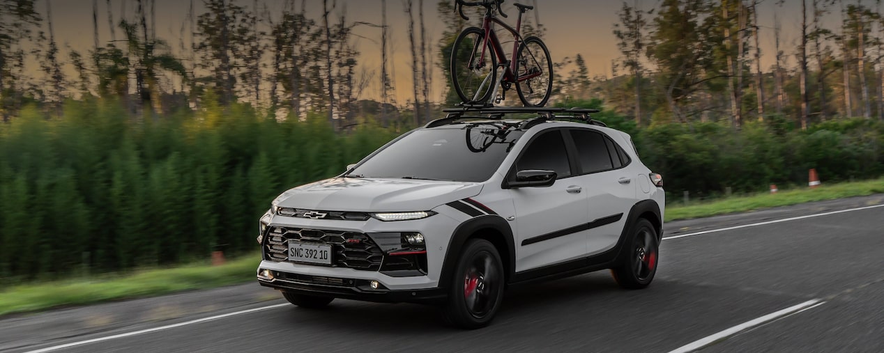 Chevrolet Sonic em movimento em uma estrada. Ao fundo vemos alguns arbustos e árvores. Em cima do veículo há uma bicicleta fixada no rack de teto. No canto superior esquerdo, o texto: "Sonic. Conforto"