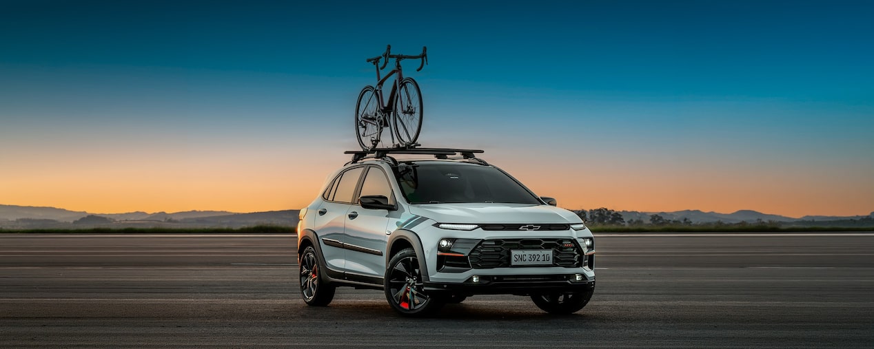 Chevrolet Sonic estacionado em uma área externa. O céu está com uma coloração levemente avermelhada de fim de tarde e o veículo está com os faróis ligados. Há uma bicicleta fixada no rack de teto do veículo. No canto superior esquerdo, o texto: Sonic. Pacotes