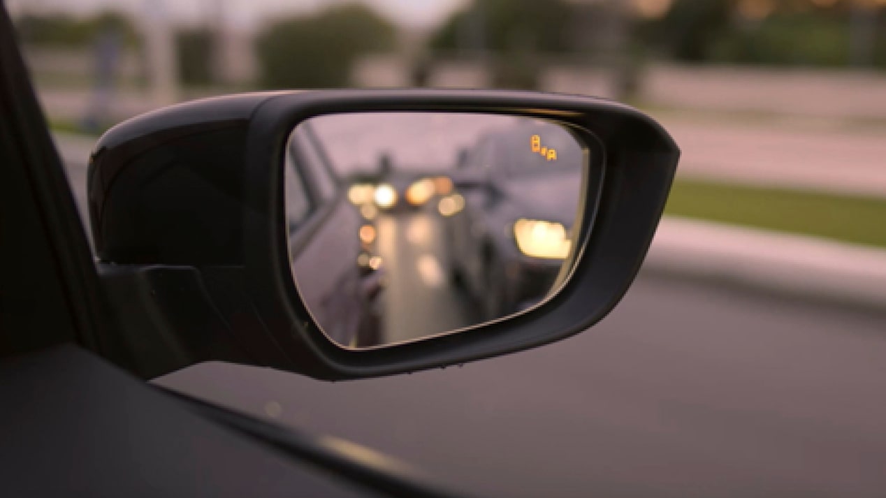 Foco no retrovisor externo direito. No espelho, temos alguns carros fora de foco e, no canto superior direito do retrovisor, vemos o aviso de ponto cego ligado.