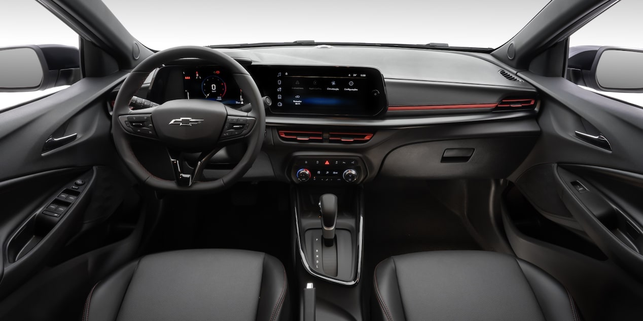 Visão em primeira pessoa do motorista. Temos o volante com o logo da Chevrolet no centro, o painel de instrumentos  e a central multimídia, câmbio automático e parte dos bancos.​