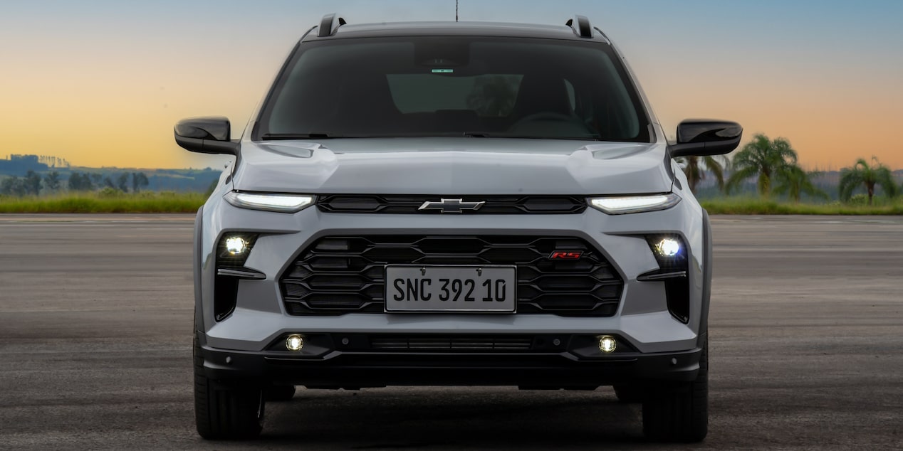 Temos a parte frontal do Chevrolet Sonic em destaque. As lanternas e faróis estão ligados. Ao fundo, temos um céu de fim de tarde.​