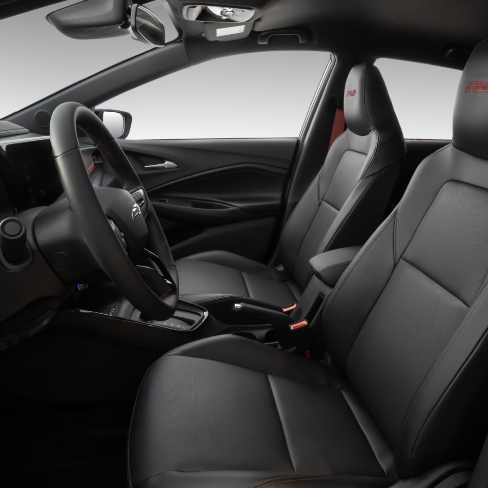 Visão em primeira pessoa do interior do carro. Temos em quadro o volante e o banco do motorista e passageiro.