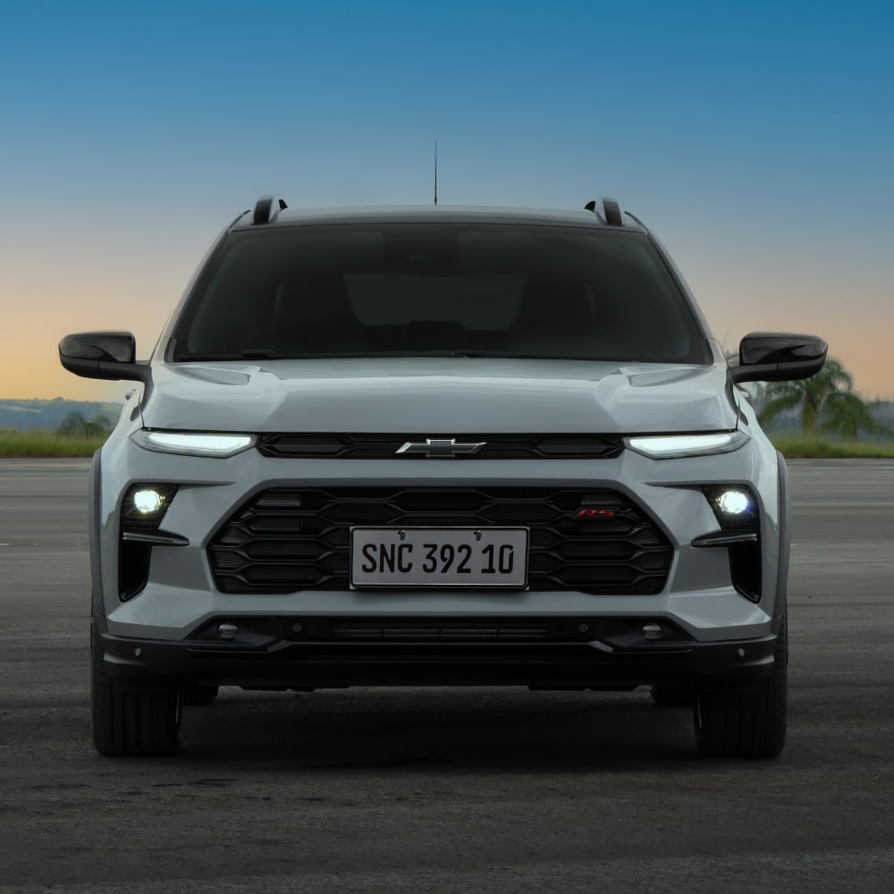 Temos a parte frontal do Chevrolet Sonic em destaque. As lanternas e faróis estão ligados. Ao fundo, temos um céu de fim de tarde.
