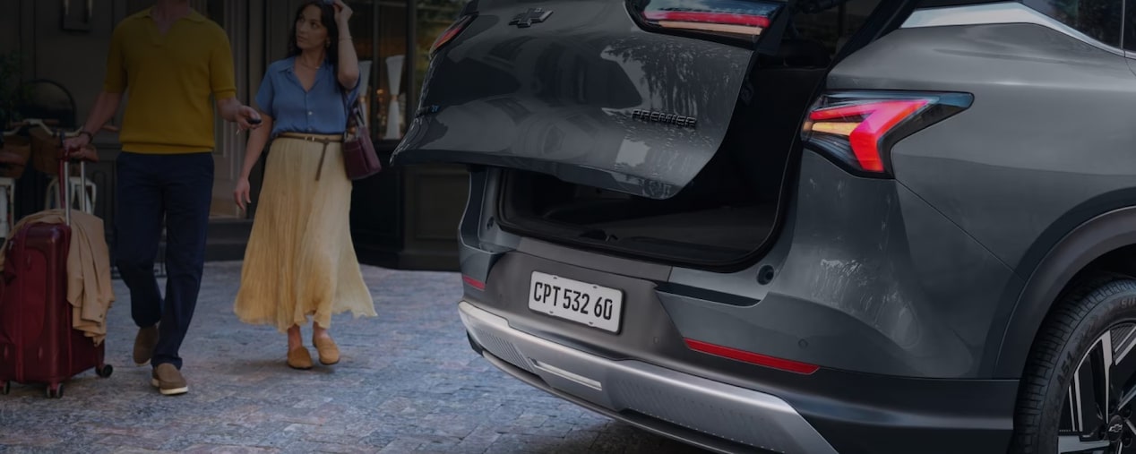 Chevrolet Captiva EV em foto ambientada. estacionado, com a tampa do porta-malas aberta, enquanto um casal se aproxima. Esta imagem ilustra a sessão de gerenciamento de carga da linha de acessórios originais Chevrolet.