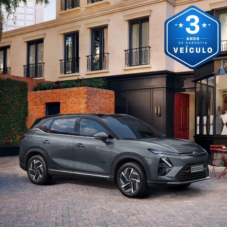 Imagem de um Captiva EV estacionado em frente a um café com mesas externas, em uma rua de paralelepípedos cercada por prédios elegantes com um selo azul indicando 3 anos de garantia do veículo.