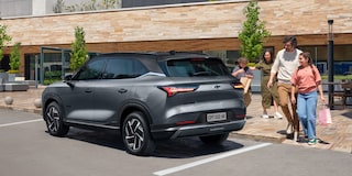 Família caminhando sorridente em direção a um SUV Chevrolet Captiva EV estacionado em frente a um prédio moderno com fachada de pedra.