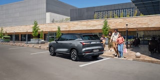 Família caminhando sorridente em direção a um SUV Chevrolet Captiva EV estacionado em frente a um prédio moderno com fachada de pedra.