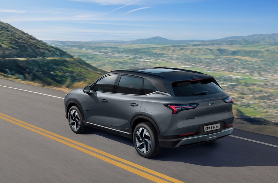 Traseira da Chevrolet Captiva EV em movimento em uma estrada sinuosa cercada por montanhas e vegetação sob céu azul.