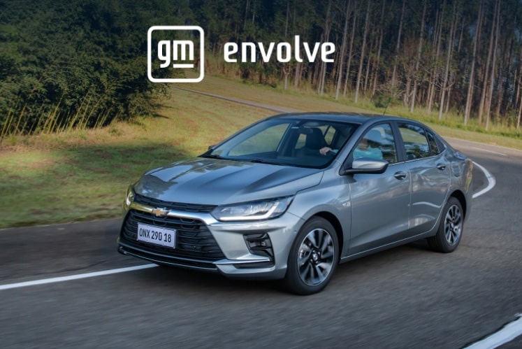Imagem do Onix Plus com logotipo GM Envolve sobreposto, em estrada dom árvores ao fundo