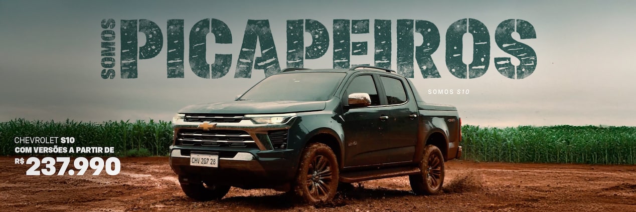 Uma picape Chevrolet S10 trafega por uma estrada de terra lamacenta ao lado de um campo, com os dizeres SOMOS PICAPEIROS e SOMOS S10 ao fundo.