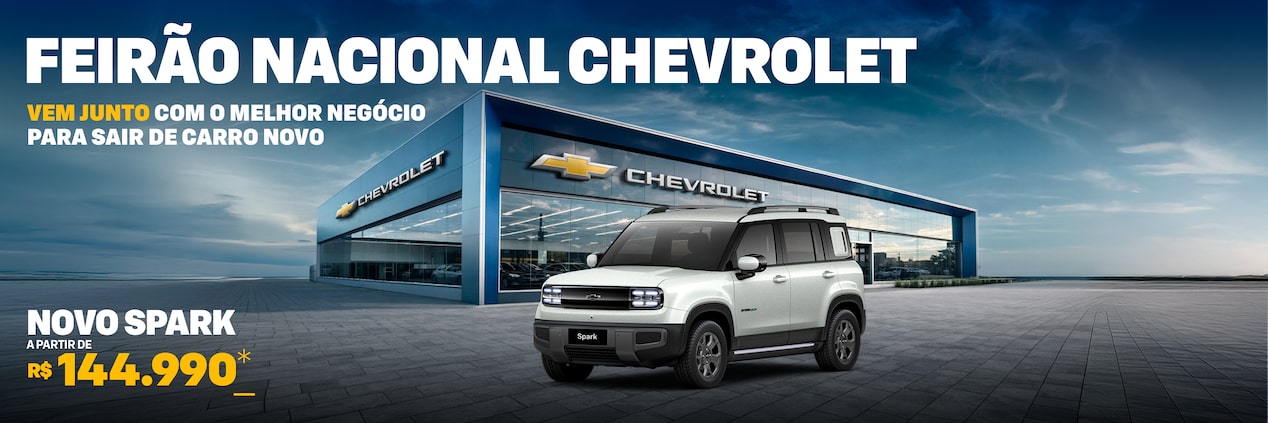 Um SUV elétrico Spark EUV branco está estacionado em frente a uma concessionária Chevrolet moderna sob um céu parcialmente nublado.