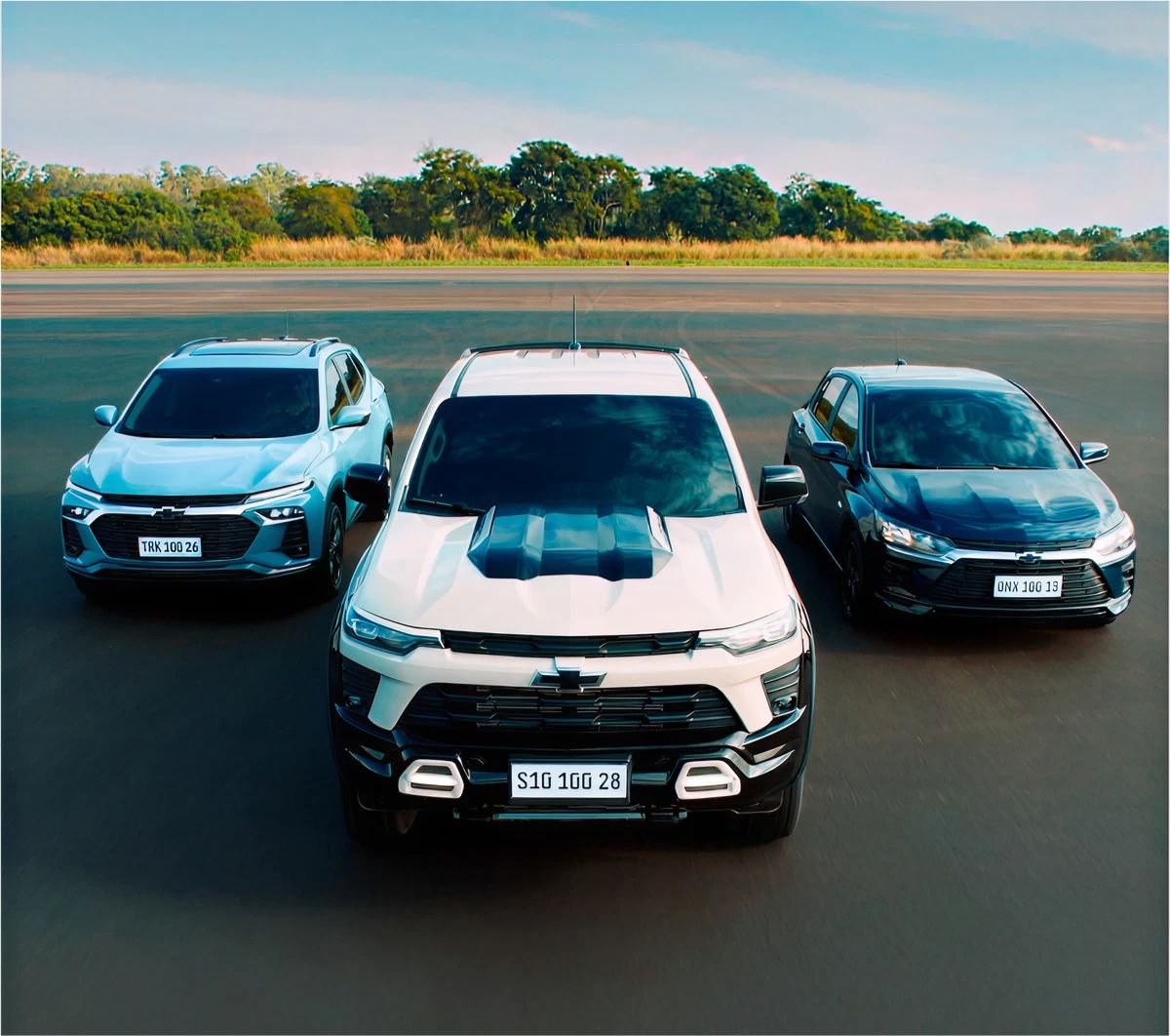 Três veículos Chevrolet alinhados em pista aberta.  S10 ao centro, Tracker à esquerda e um Onix à direita, todos em vista frontal.