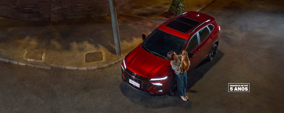 Banner Novo Chevrolet Tracker, cor vermelha, visto de cima em uma rua à noite. Uma mulher segura um cão pequeno ao lado do carro. Texto promocional: Evoluímos onde mais importa. Taxa zero e parcelas de apenas R$ 2.299 com 5 anos de garantia. Selo Garantia de até 5 anos no canto  inferior direito.