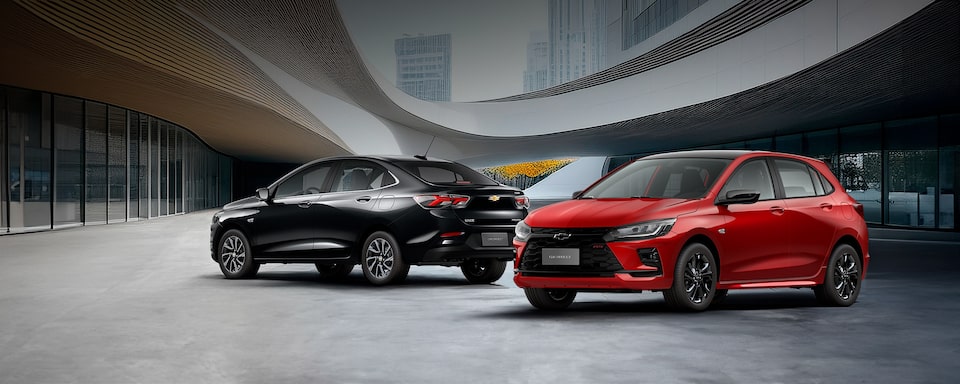 Um Onix Plus preto e um Onix  vermelho, estacionados lado a lado em frente a um prédio moderno com estrutura de vidro. Texto sobreposto: Paixão nacional: Onix e Onix Plus.