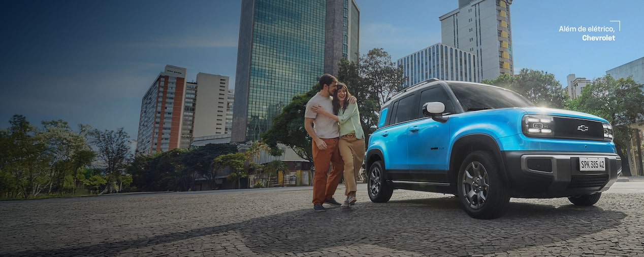 Banner de lançamento do Chevrolet Spark EV visto de frente e lateral direita, estacionado em avenida urbana com prédios altos ao fundo. À esquerda, texto promocional: Novo Chevrolet Spark – condição especial de lançamento a partir de R$ 159.990 com taxa zero em 24x. À frente do carro, um casal sorridente se abraça. No canto superior direito, slogan Além de elétrico, Chevrolet.