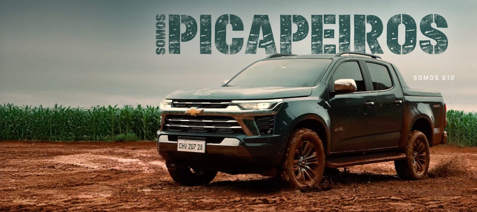 Uma picape Chevrolet S10 trafega por uma estrada de terra lamacenta ao lado de um campo, com os dizeres SOMOS PICAPEIROS e SOMOS S10 ao fundo.